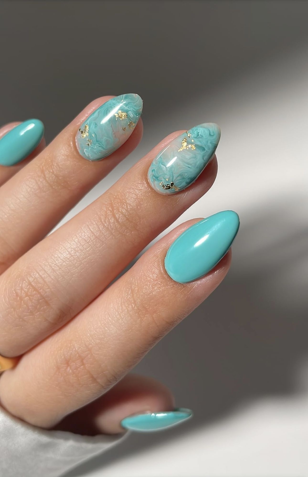 Close-up of a hand with aqua-colored almond-shaped nails. Two nails feature a marbled design with gold flecks, while others…