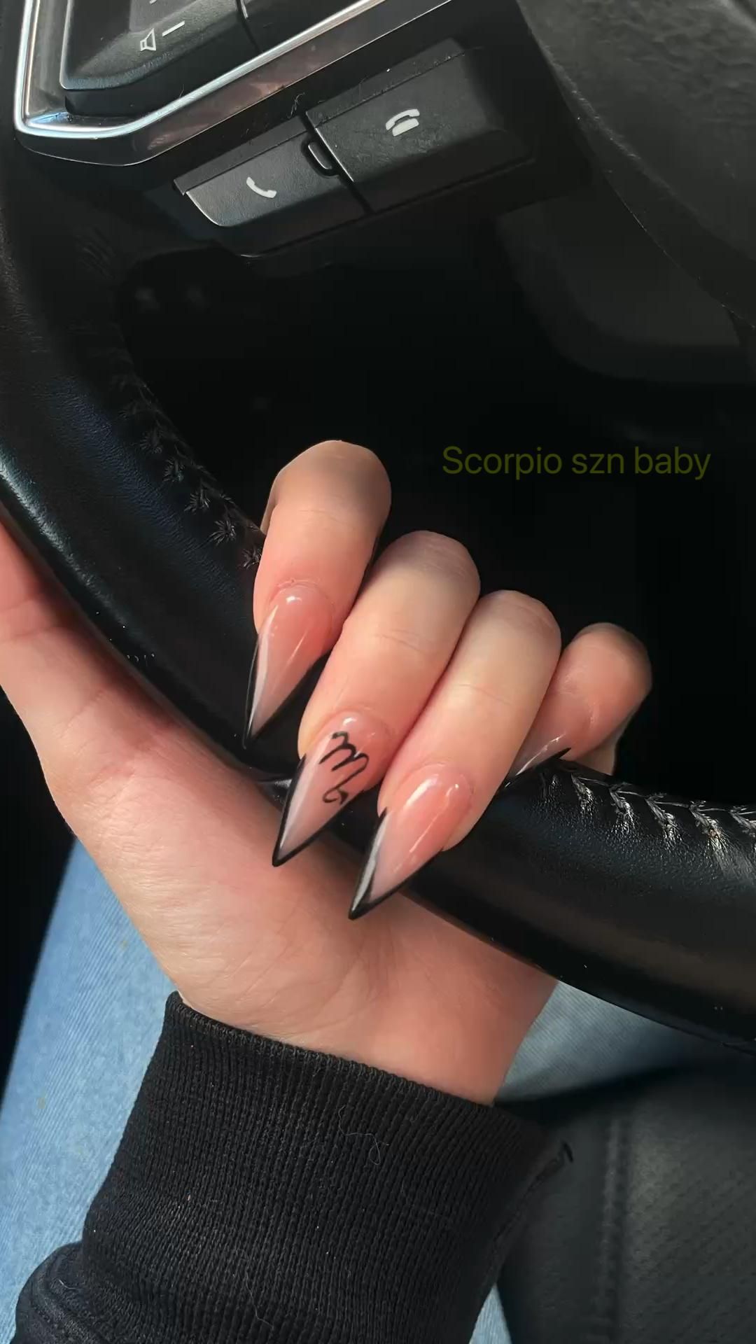 Hand with stiletto nails featuring black tips and the Scorpio symbol on one nail, holding a steering wheel.