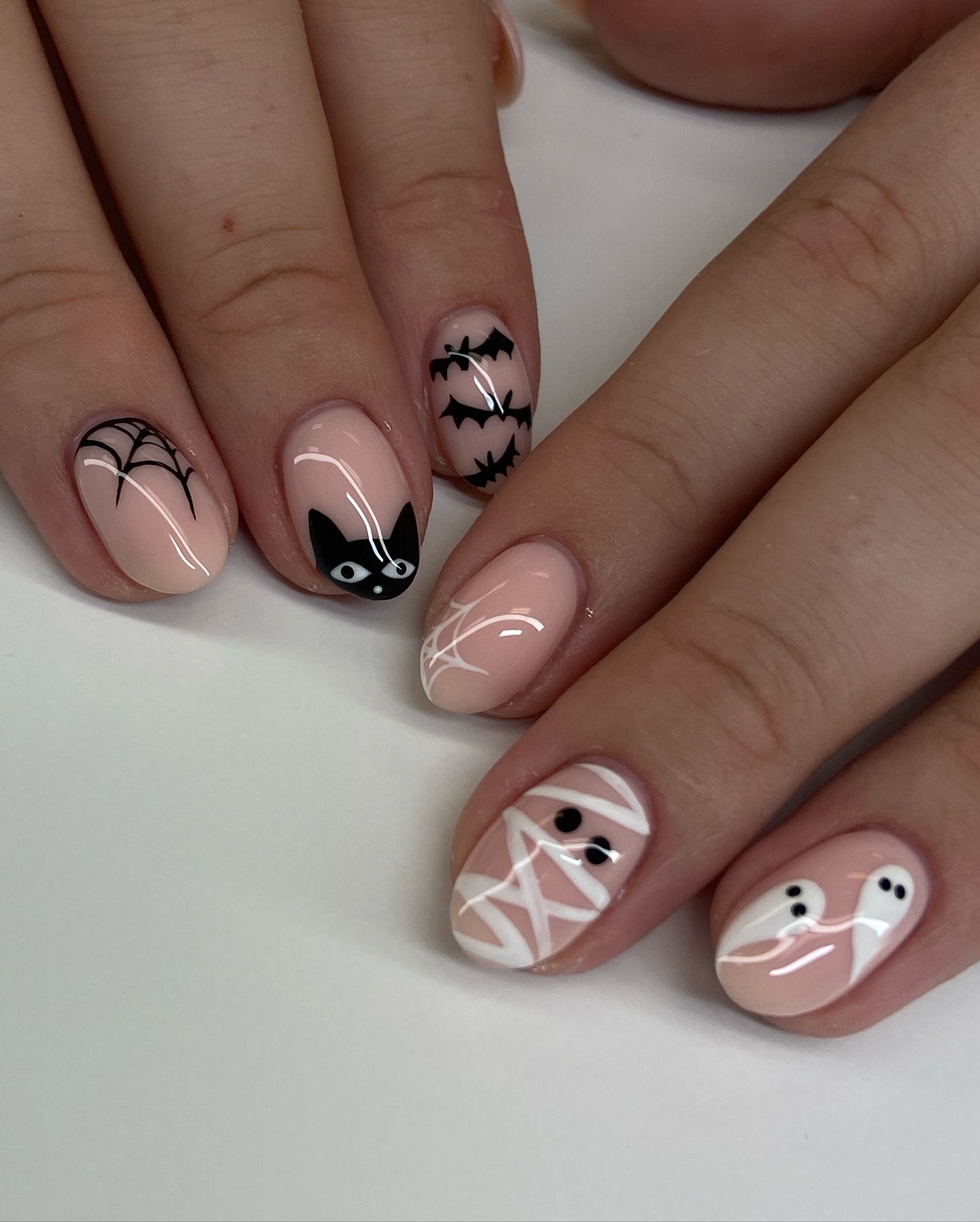 Close-up of manicured short nails with various Halloween designs: spiderweb, black cat, bats, white abstract lines, mummy…
