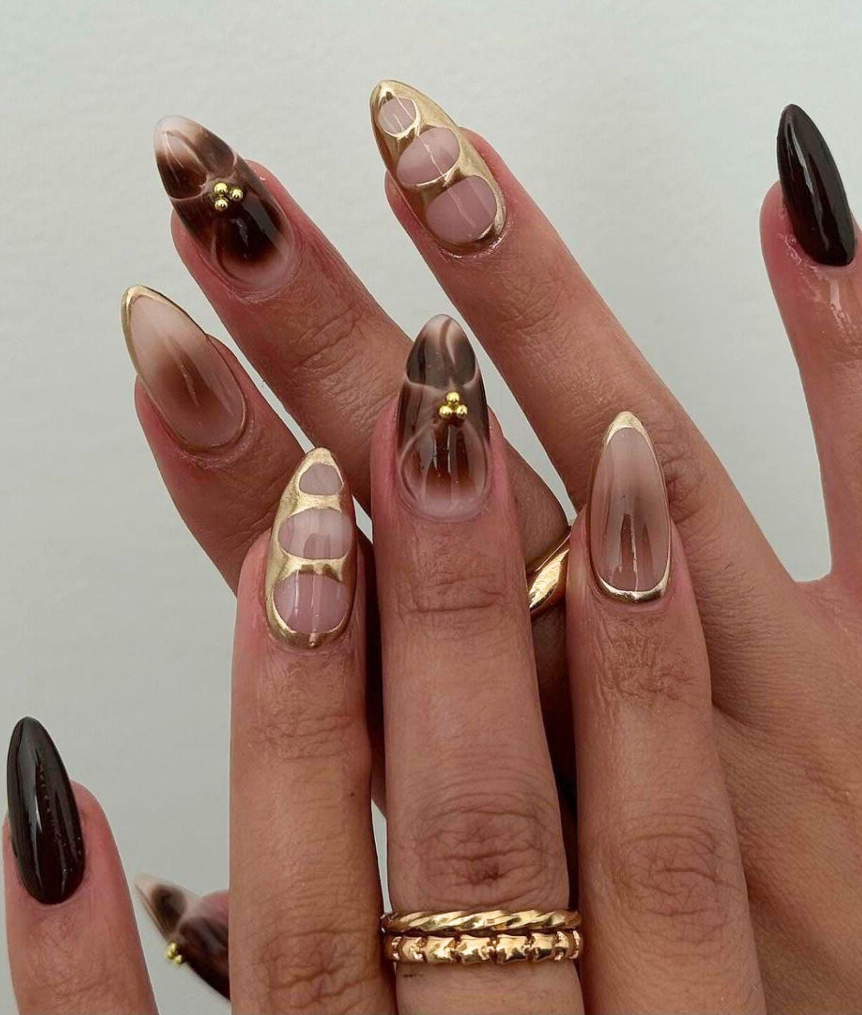 Close up of manicured hands with almond-shaped acrylic nails. Some nails are a brown ombré, others are solid brown, and some…