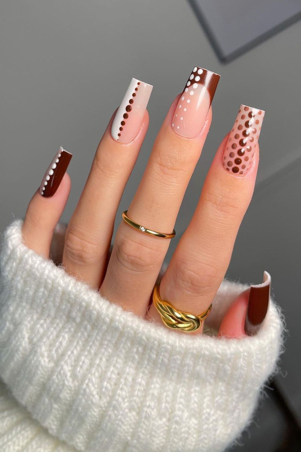 Hand with long, square-tipped acrylic nails in shades of brown, white, and nude, featuring various dot patterns.