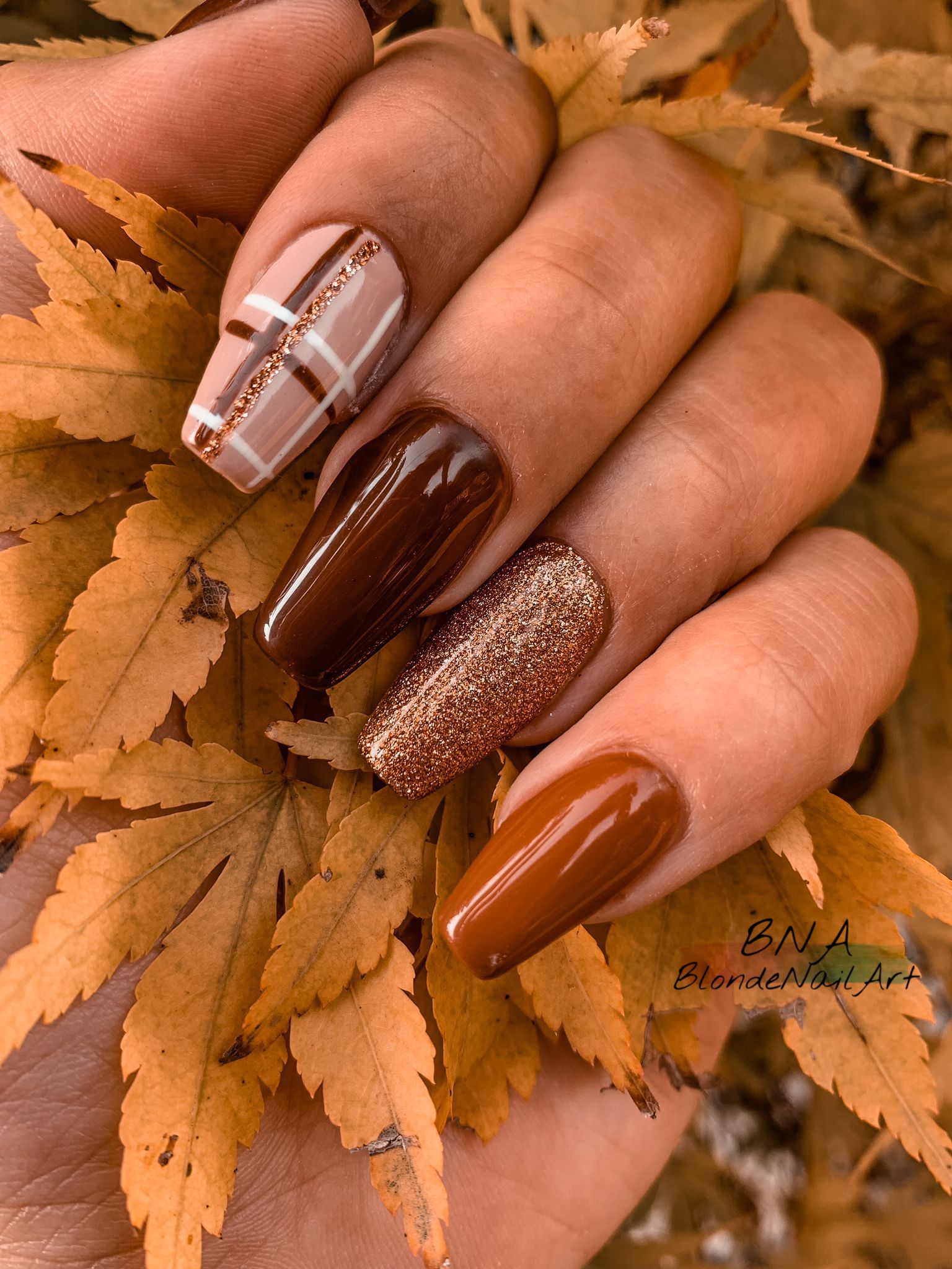 Hand with coffin-shaped nails featuring a plaid pattern, a solid dark brown, a glittery gold, and a solid reddish brown, all…