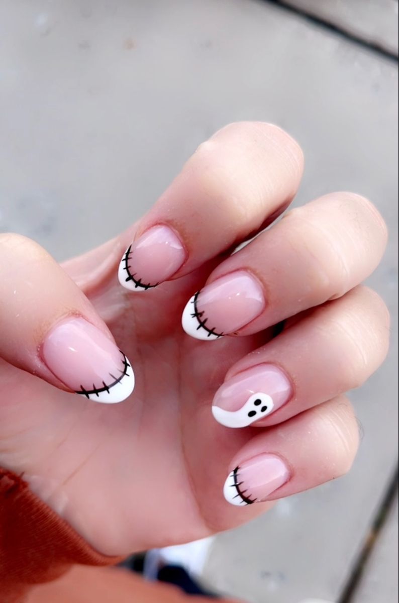 Close-up of a hand with short, round-shaped nails. The nails feature white French tips with black stitch detailing, and one…