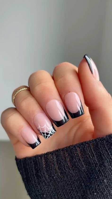 Close-up of a hand with square-shaped nails, featuring black French tips. One nail has a white spiderweb design.