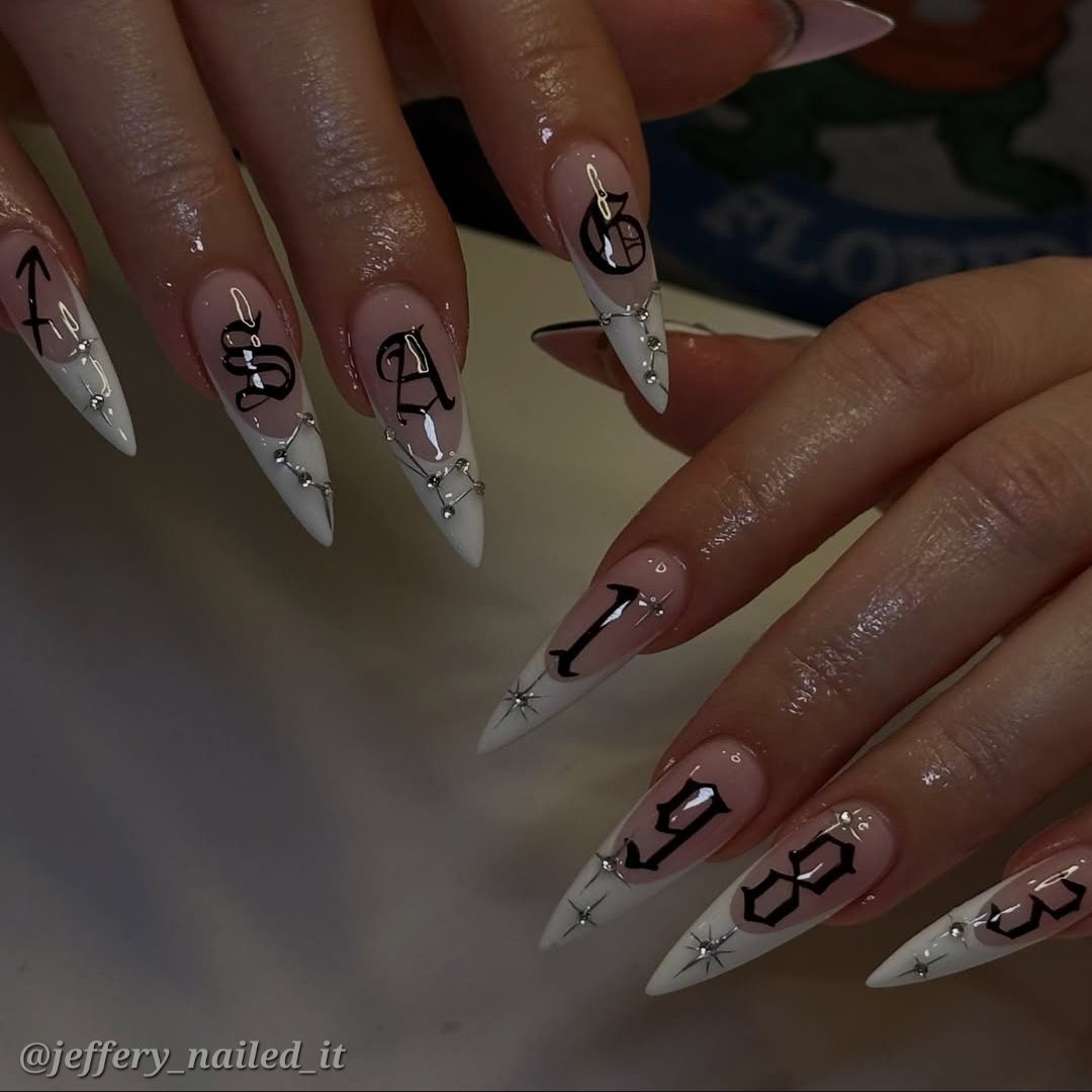Long stiletto nails with white tips, a nude base, and black gothic letters forming a name, connected by silver chains and…