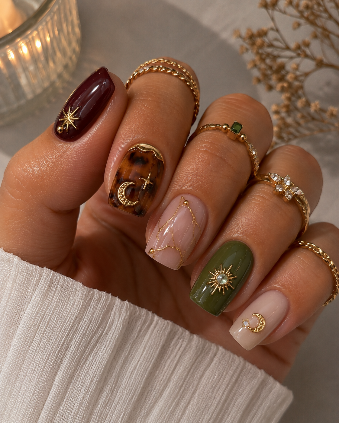 Close-up of a hand with nails painted in varying dark red, tortoise shell, olive green, and nude with gold celestial charms…