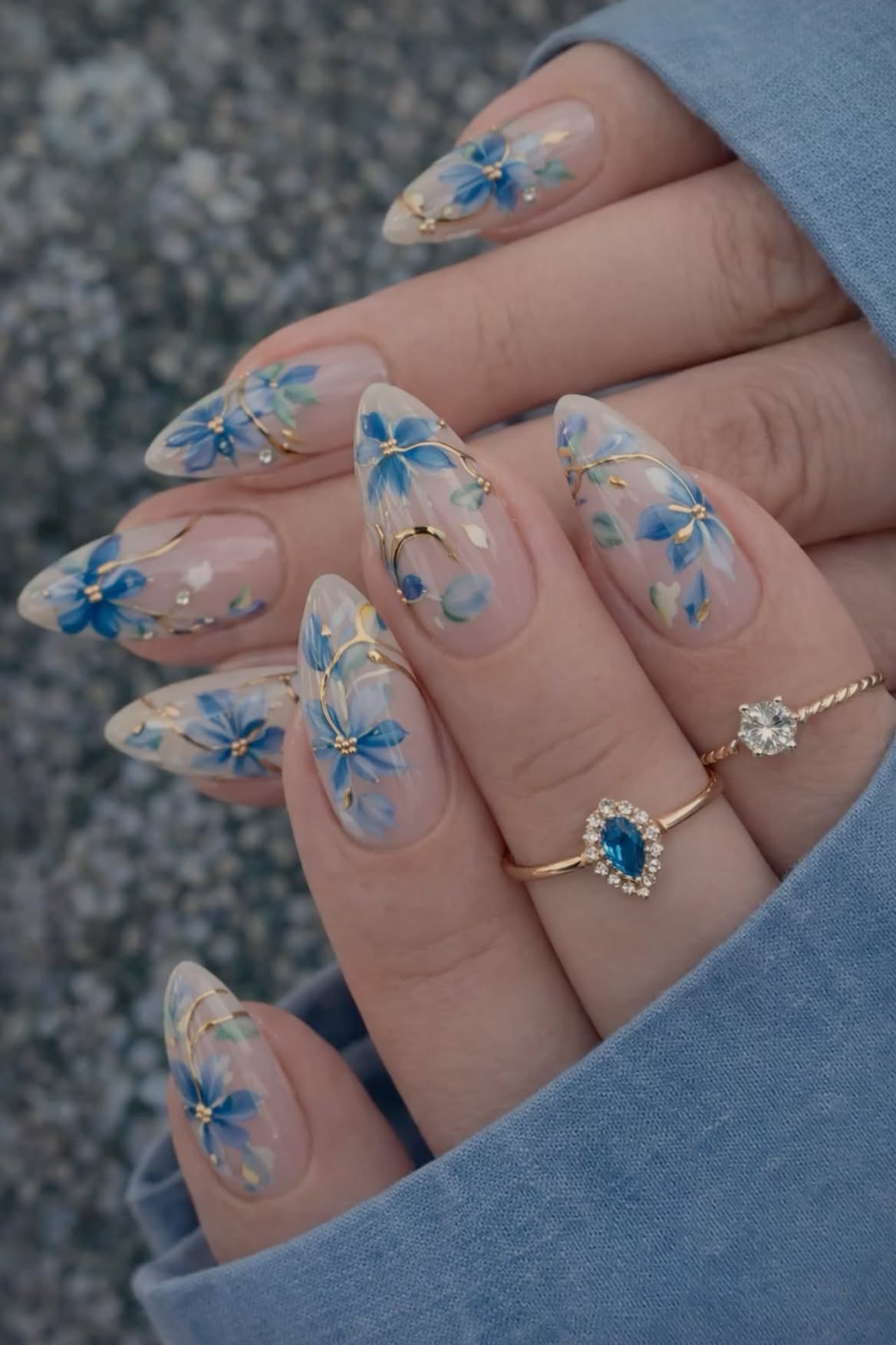 Pointed almond nails adorned with delicate blue floral patterns, gold accents, and subtle glitter details.