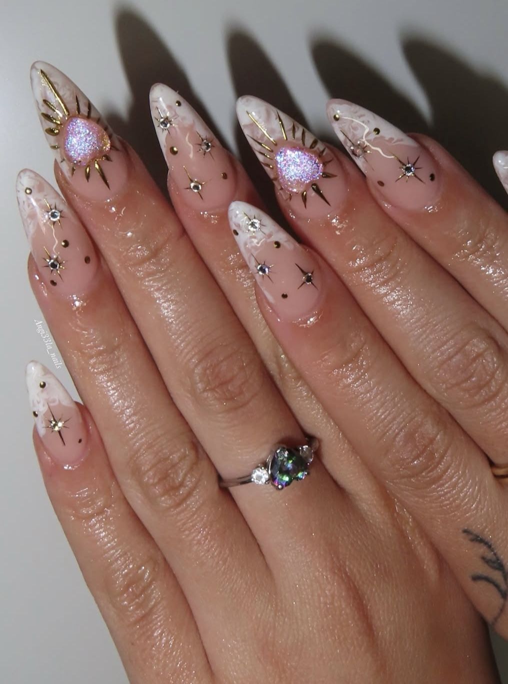 Close-up of almond-shaped stiletto nails featuring a clear base with white tips, gold accents, small gems, and starburst…