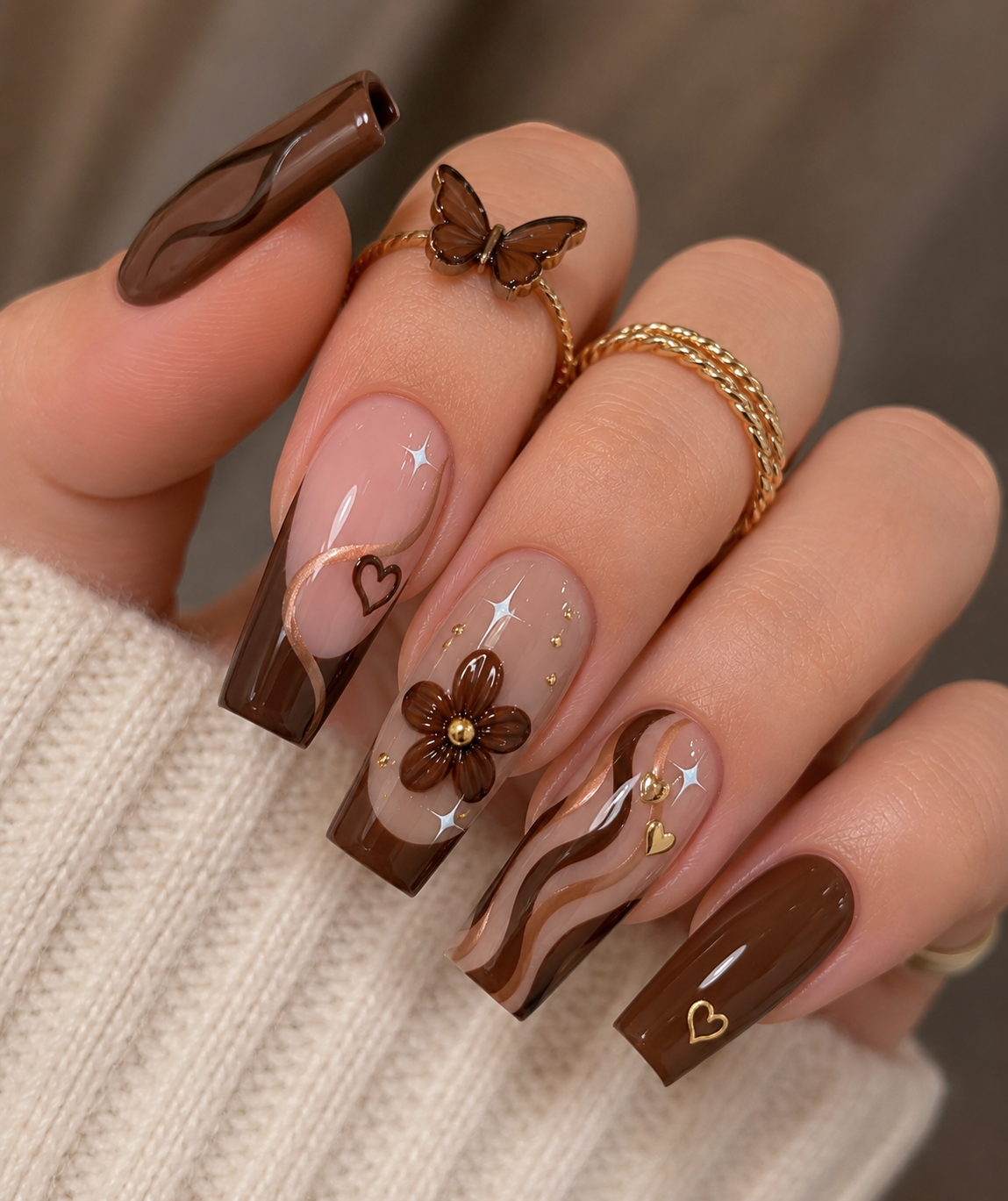 Close up of a hand with long coffin-shaped nails painted in shades of brown and nude with gold accents.