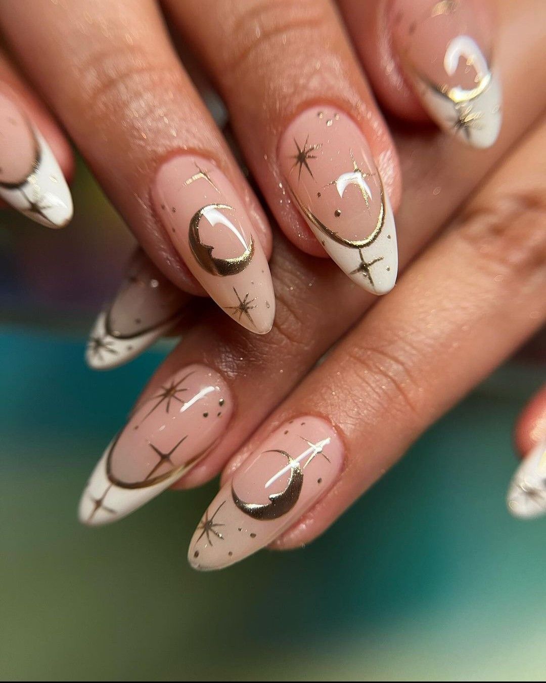 Close-up of manicured almond-shaped nails with nude base, white French tips, and gold celestial accents including moons and…