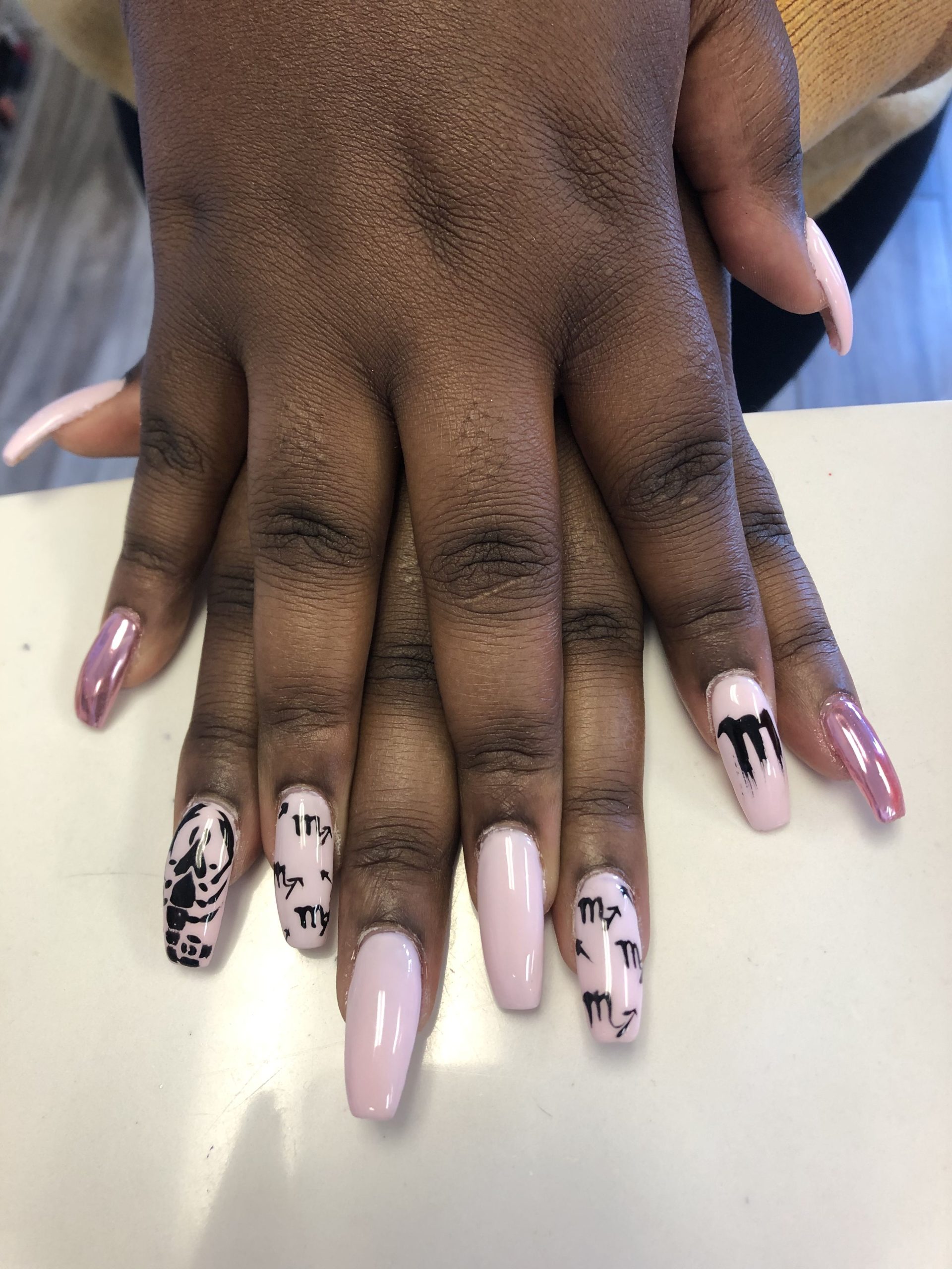 Hands with light pink and metallic pink nails. Some nails feature black Scorpio zodiac symbols and a dripping design.