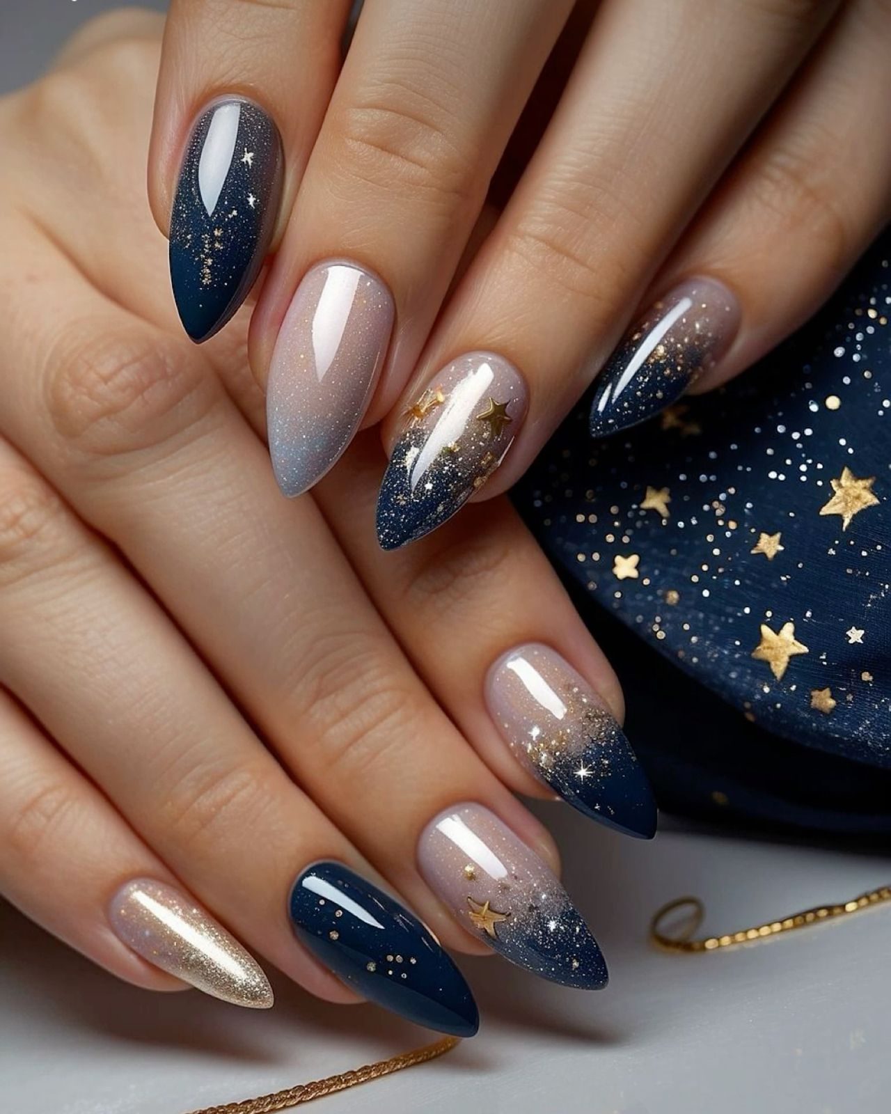 Almond-shaped nails with a glitter ombre design, fading from navy blue to nude, accented with gold stars and glitter.