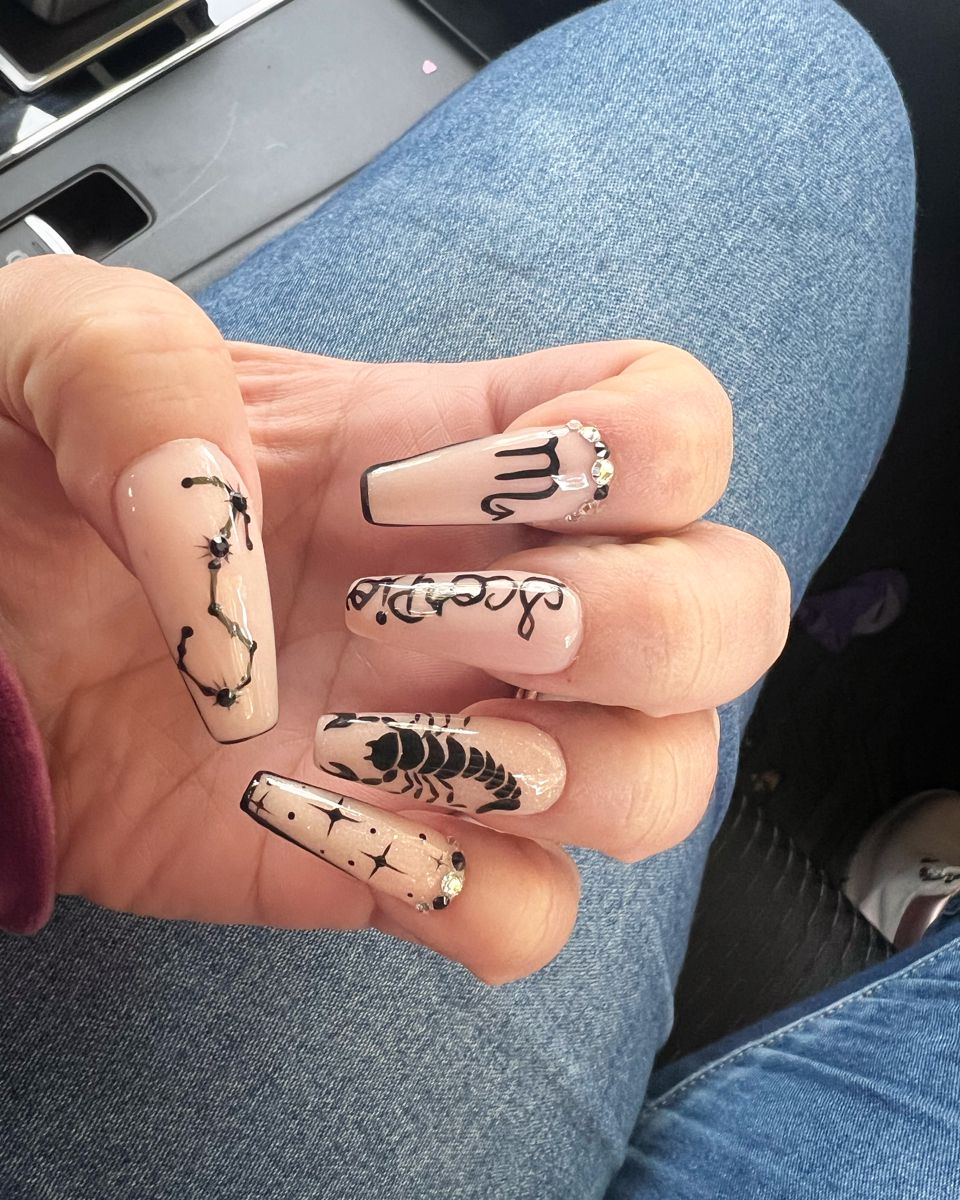 Five long nails with a light neutral base. Each nail features a different black design, including a constellation, the word…