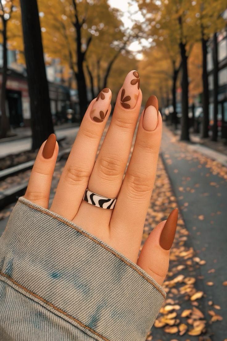 Hand with coffin nails featuring brown solid and leaf-patterned designs, wearing a black and white ring.