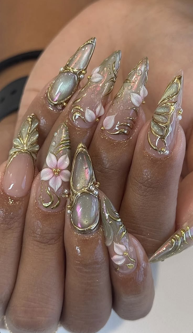 Close-up of a hand with very long, ornate stiletto nails. The nails feature 3D floral designs, gold accents, and pearlescent…