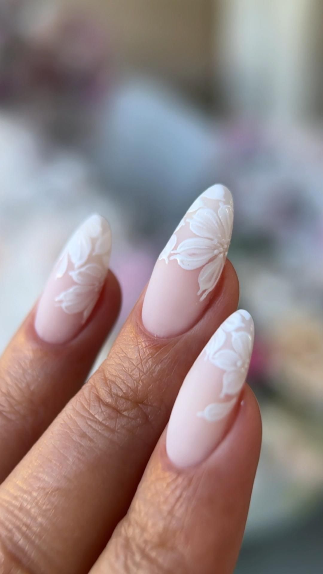 Close-up of three almond-shaped nails with a soft nude base and white floral designs at the tips.