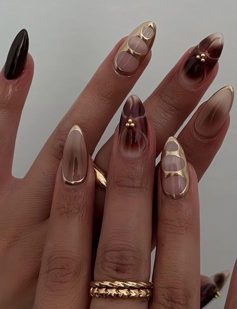 Close up of manicured hands with long almond-shaped nails. The nails are transparent brown and black with gold tips and…