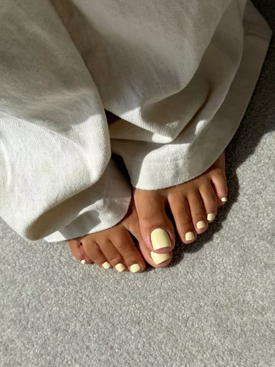 Close-up of feet with light yellow polished toenails, peeking out from under white pants.