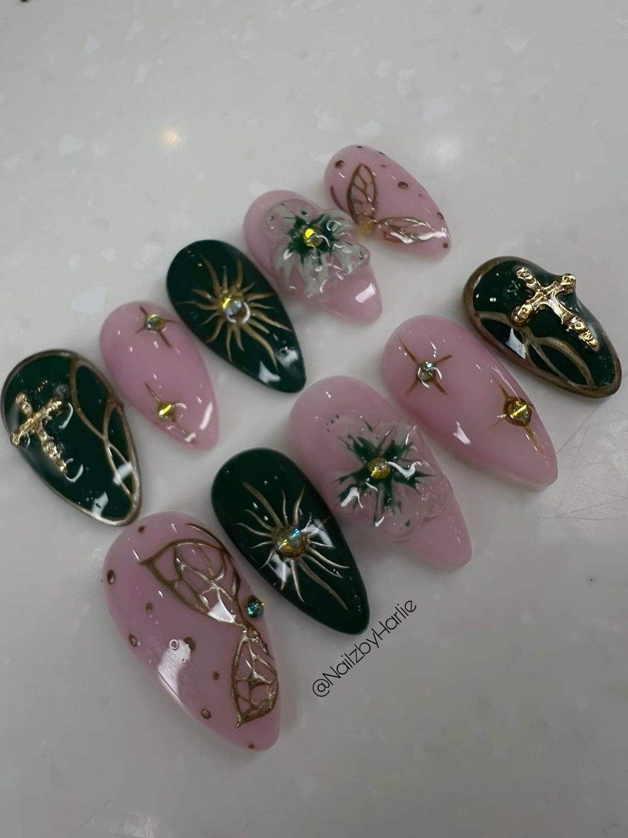 Several detached, almond-shaped press-on nails in varying shades of dark green and light pink, some with gold astrological…
