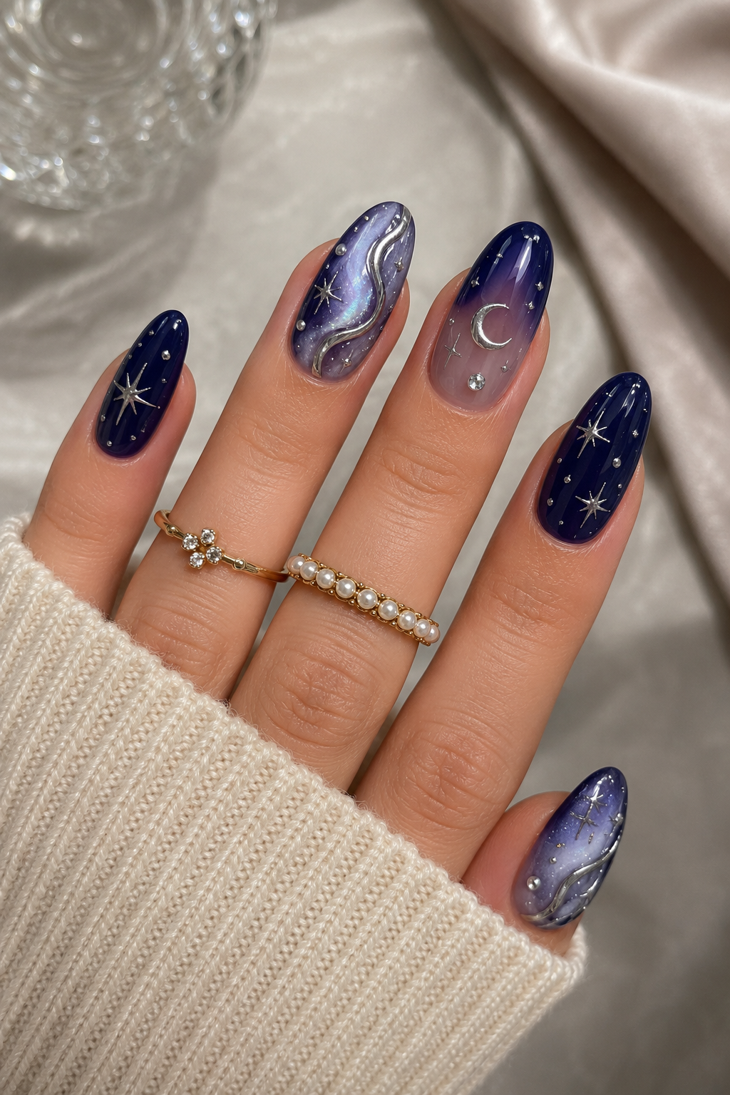 Hand with almond-shaped nails featuring deep blue and purple celestial designs with metallic silver stars and swirls…