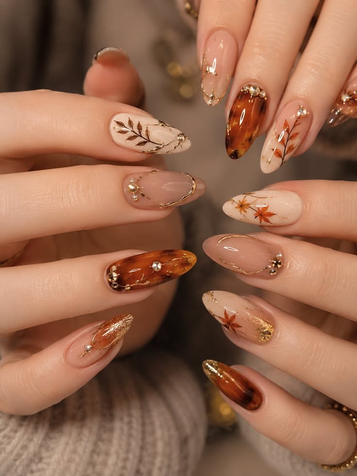 Close-up of two hands with manicured almond-shaped acrylic nails, featuring fall-themed leaf designs and jeweled accents.