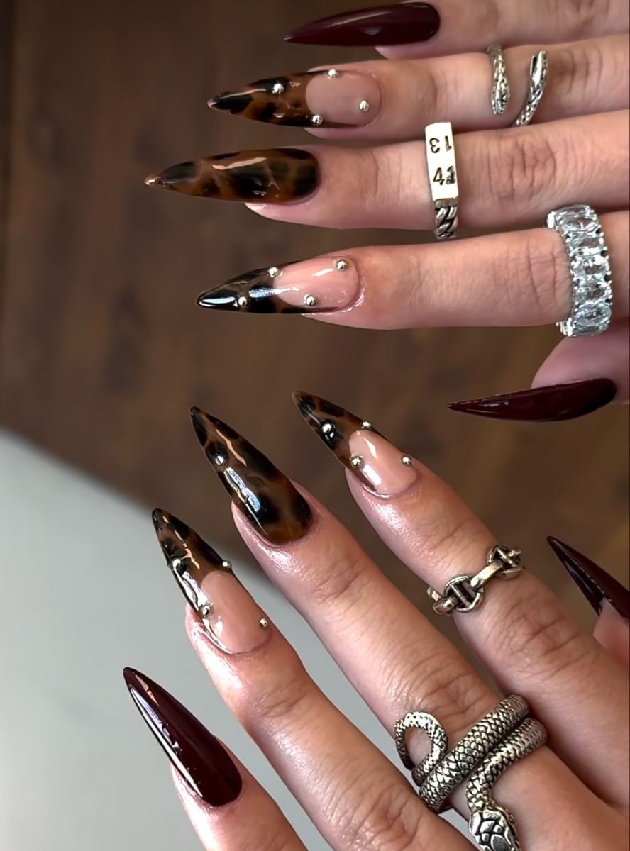 Pointed nails featuring a mix of deep berry red and tortoiseshell patterns with nude bases, accented by small silver studs.