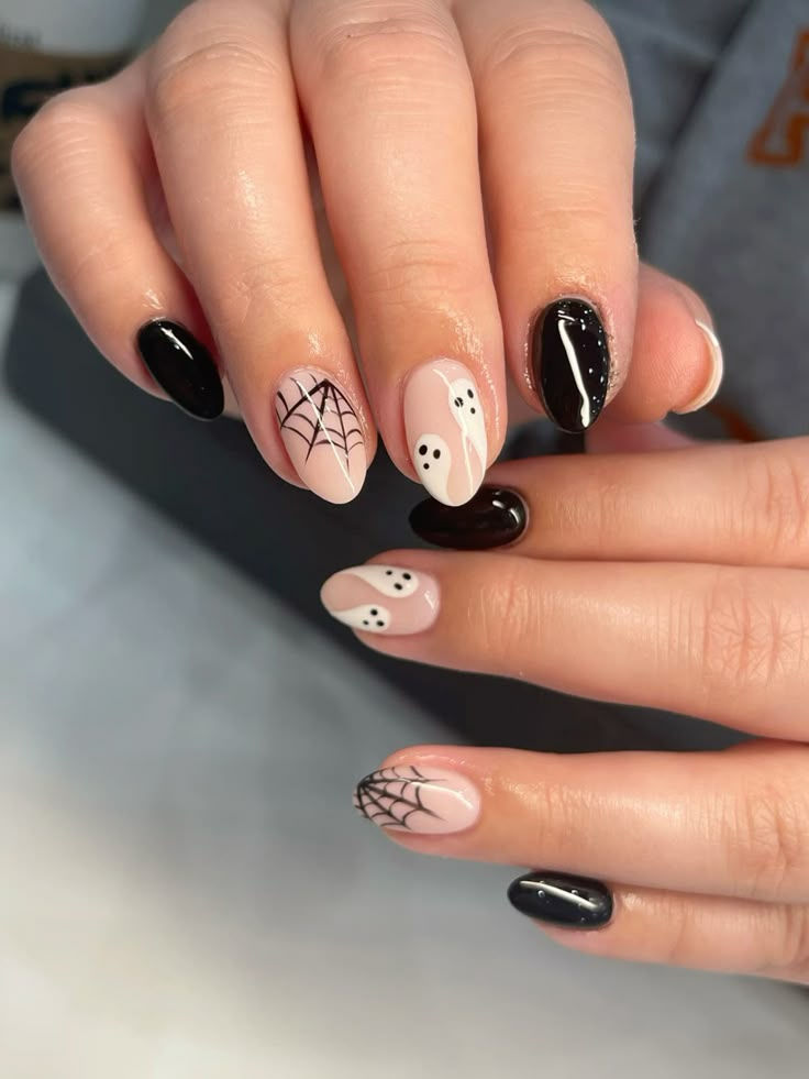 Close up of manicured hands with Halloween themed nails. Some nails are solid black, others are nude with ghost or spiderweb…