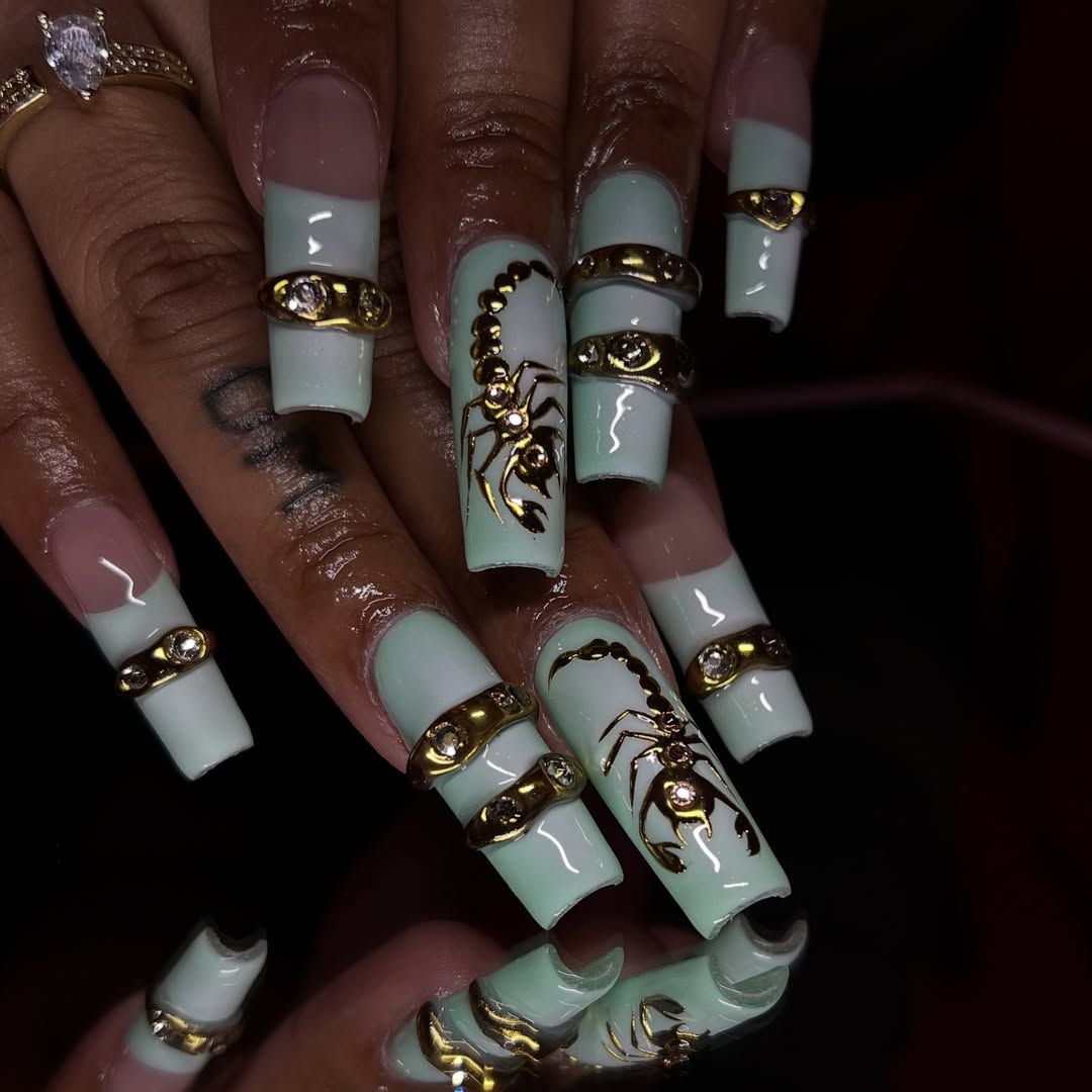 Long artificial nails with mint green tips and clear bases. Gold scorpion designs appear on some nails, while others feature…
