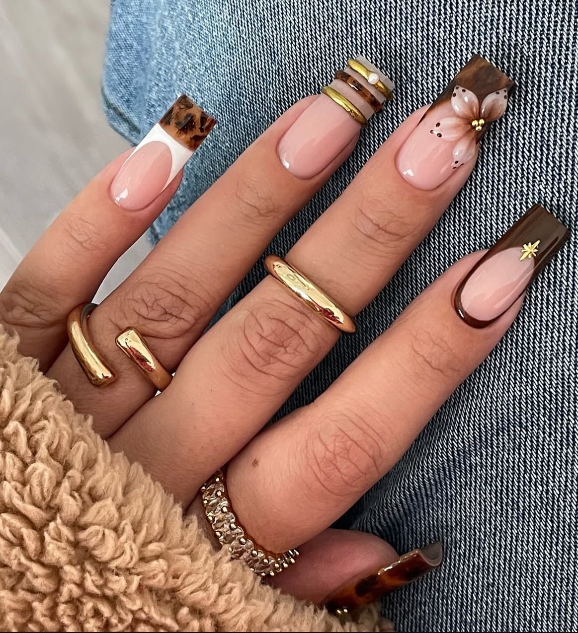 Close up of a hand with five long coffin acrylic nails featuring varying warm-toned designs against a denim background.