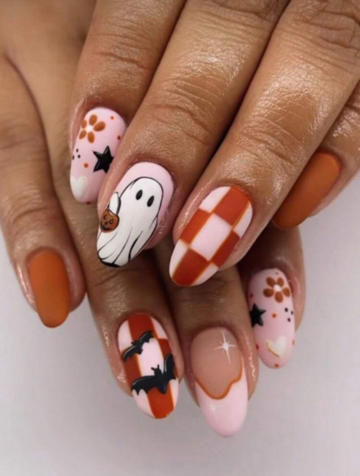 Close-up of a hand with various Halloween-themed short nail designs, including a ghost, orange checkerboard, and bats.