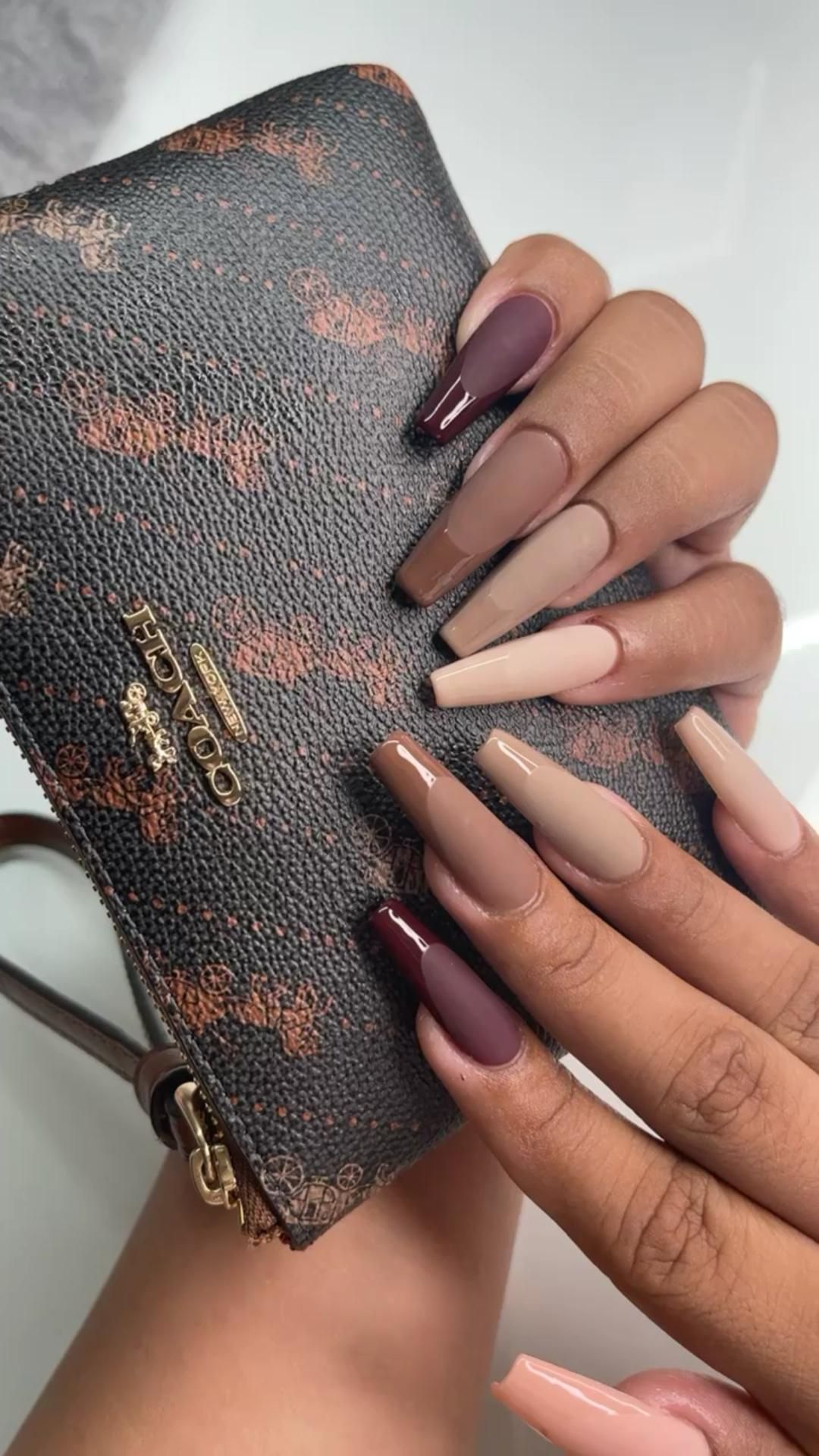 A hand displays long coffin-shaped acrylic nails in varying shades of brown, beige, and burgundy.