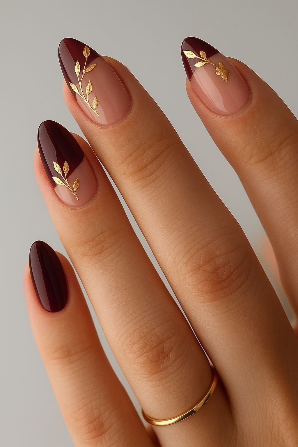Close up of a hand with almond-shaped nails featuring burgundy French tips over a nude base, adorned with intricate gold…