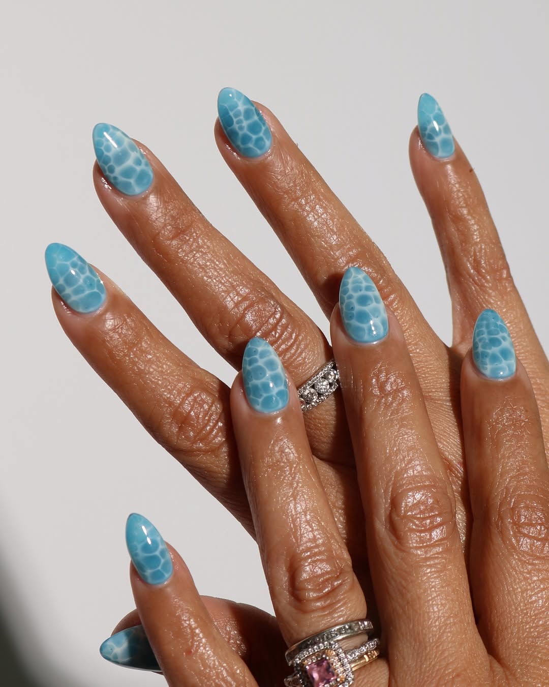 Almond-shaped aqua nails on a dark-skinned hand, featuring a distinct, irregular white pattern that resembles turtle shell…