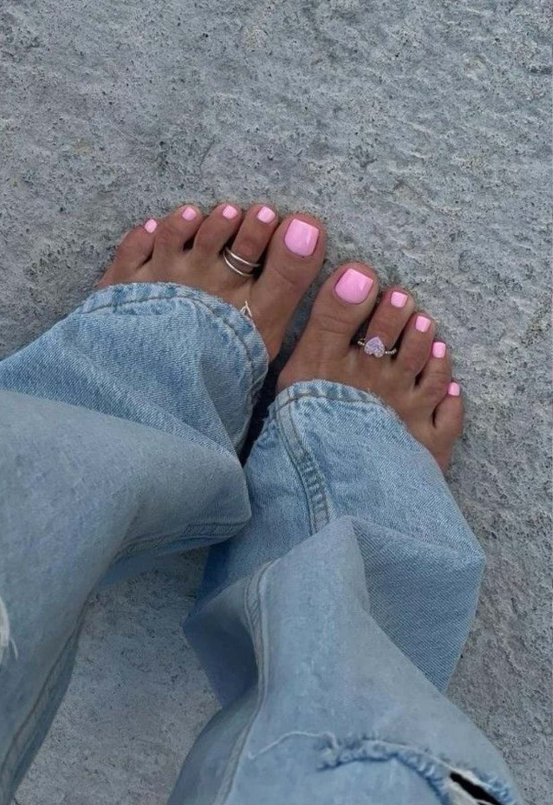 Feet with bright pink pedicure and two toe rings, wearing light blue jeans, on a gray speckled surface.