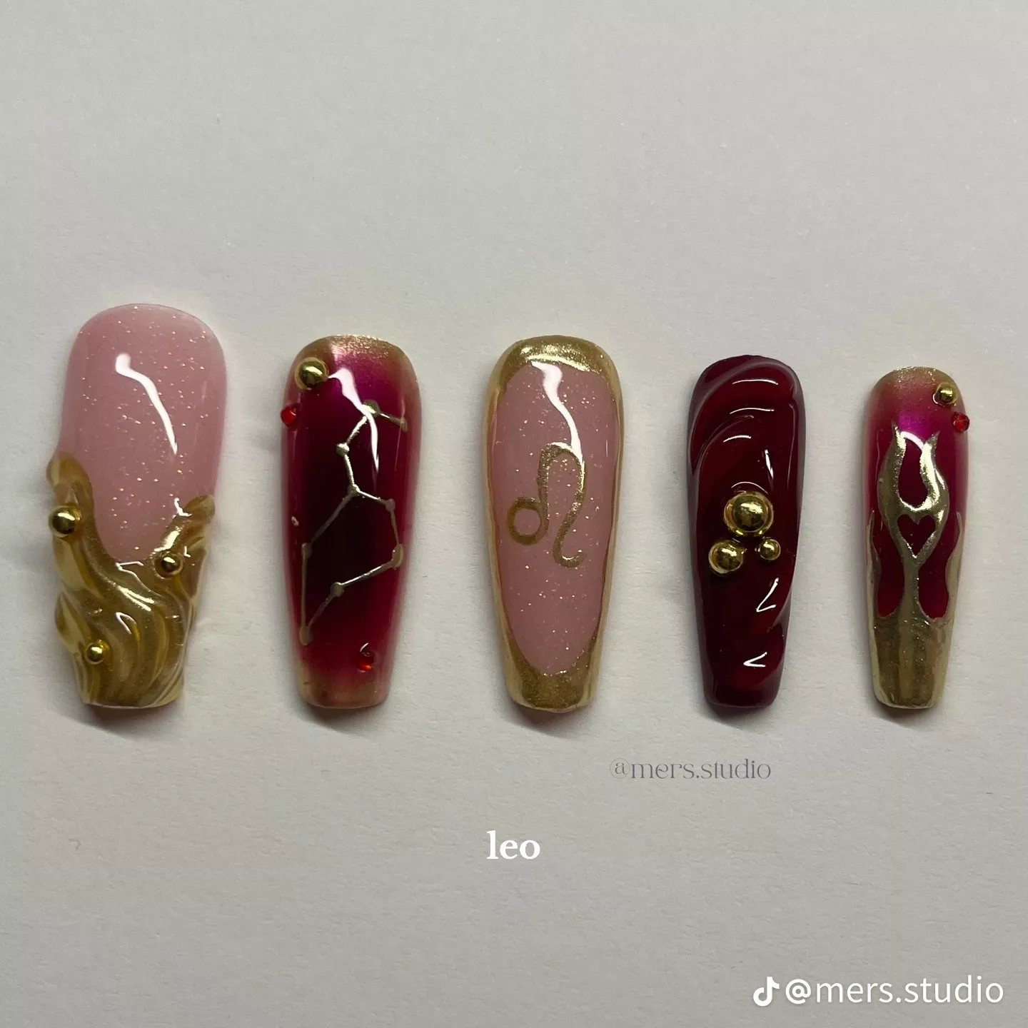Five decorated press-on nails in shades of pink, red, and gold, with metallic accents and zodiac-inspired designs.