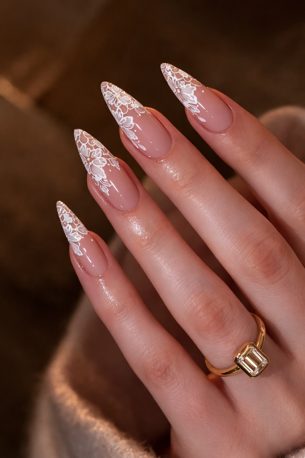 Hand with long almond-shaped nails featuring white lace-like floral designs over a nude base, wearing a gold ring with a…