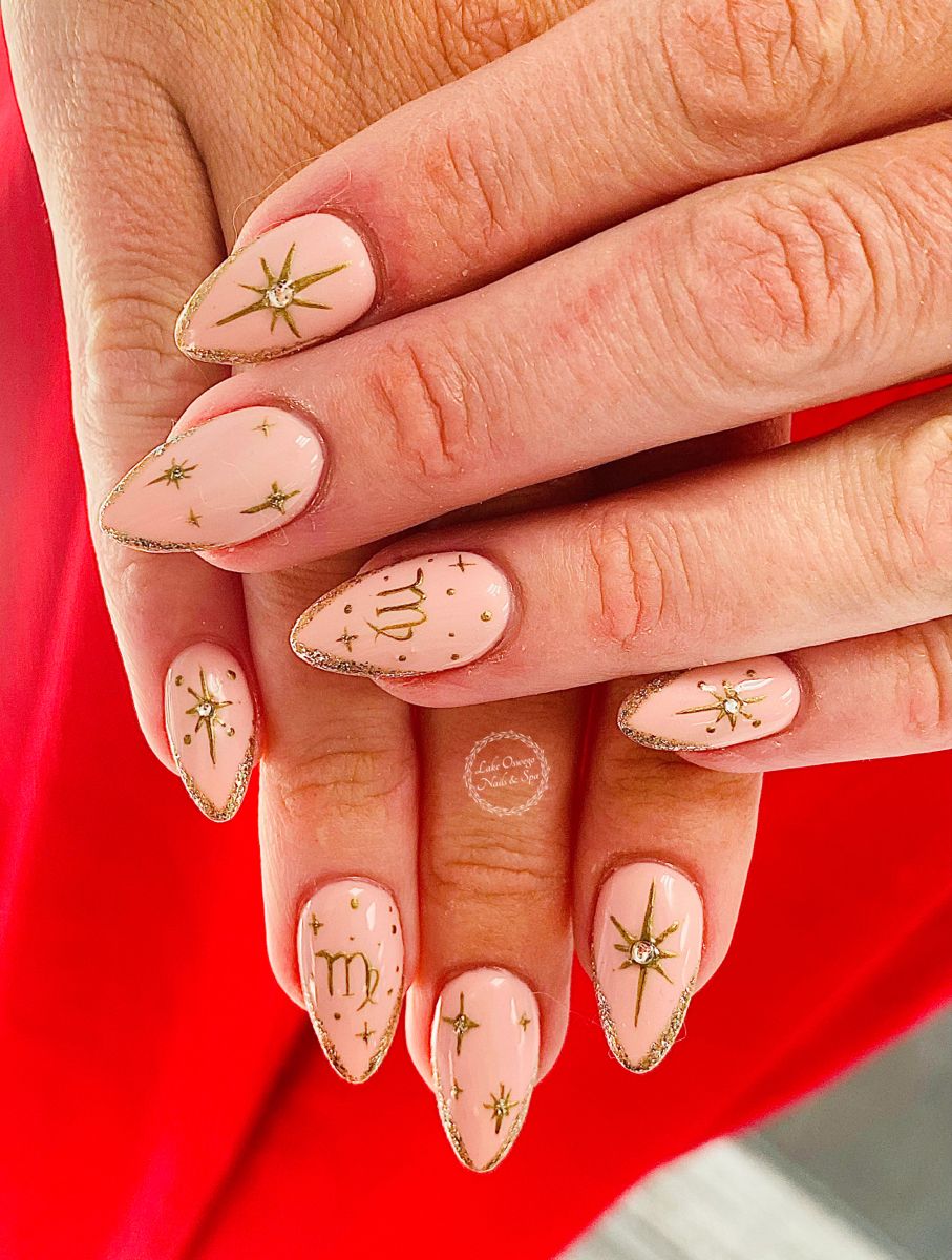 Almond-shaped, pale pink nails with gold glitter tips, gold starburst designs, and the Virgo zodiac symbol on two nails.