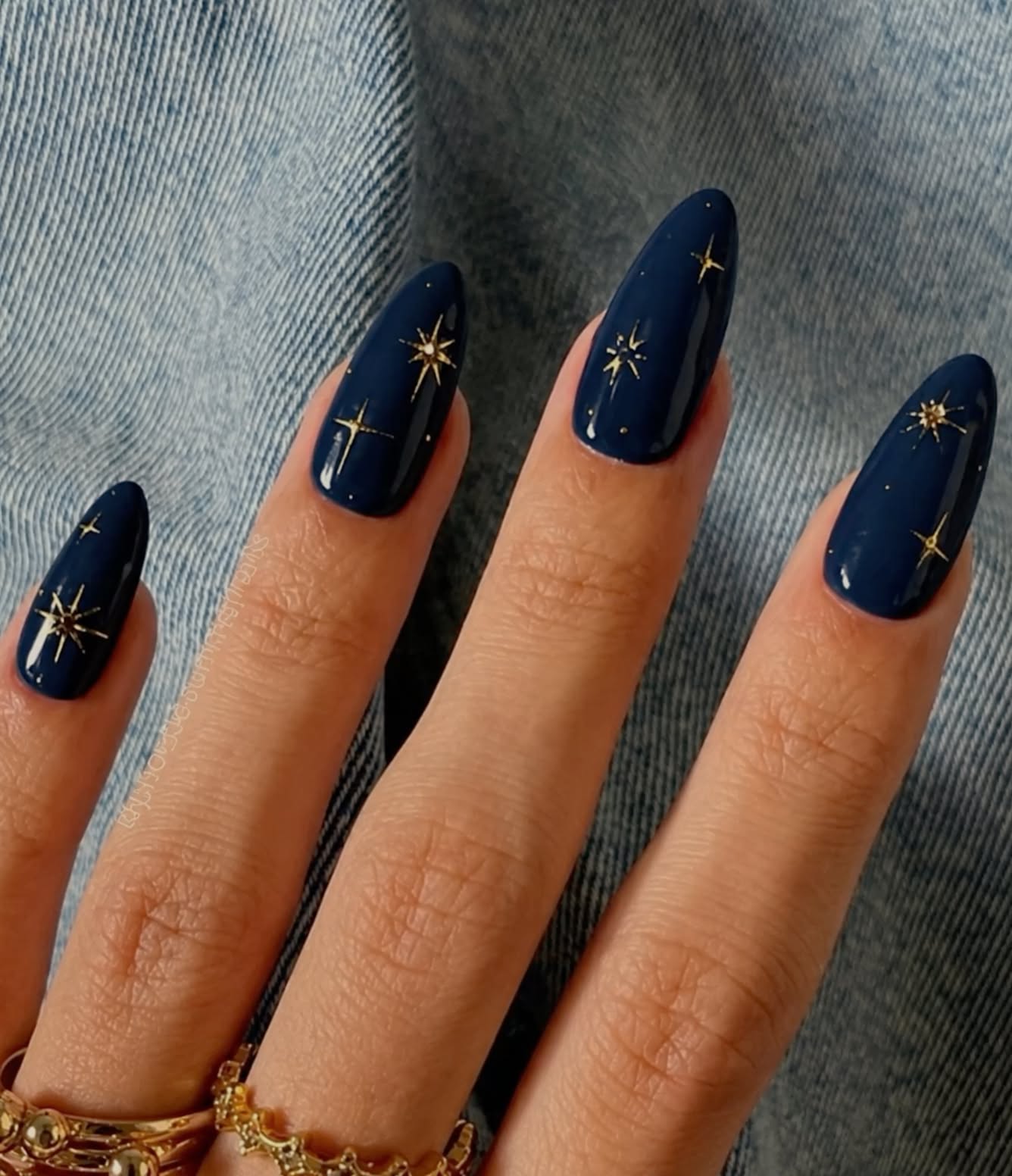 Dark blue almond-shaped nails with gold star and dot designs on four fingers, worn with gold rings.