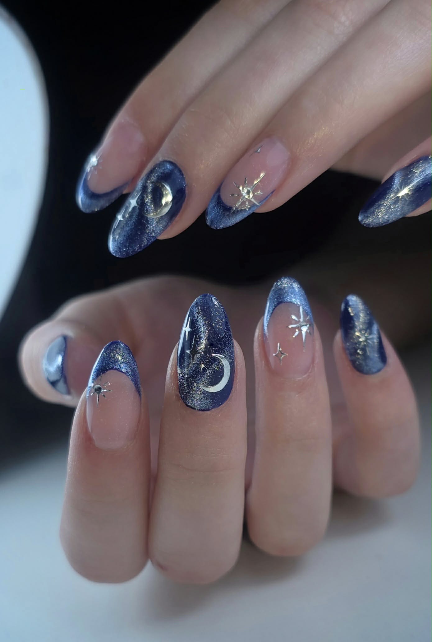 Pointed oval nails in deep blue shimmer, some with a clear base, adorned with silver moons and star accents.