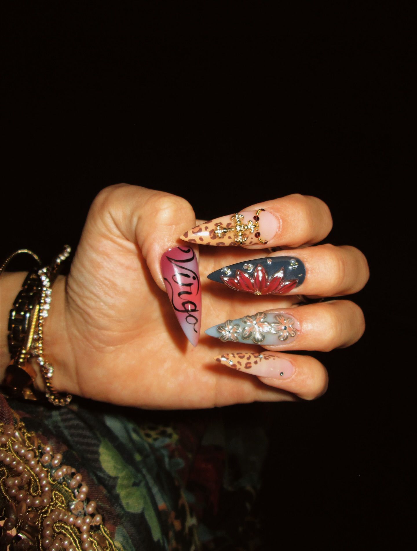 Hand with stiletto nails, one with "Virgo" text, others with leopard print, floral designs, and gemstone accents.