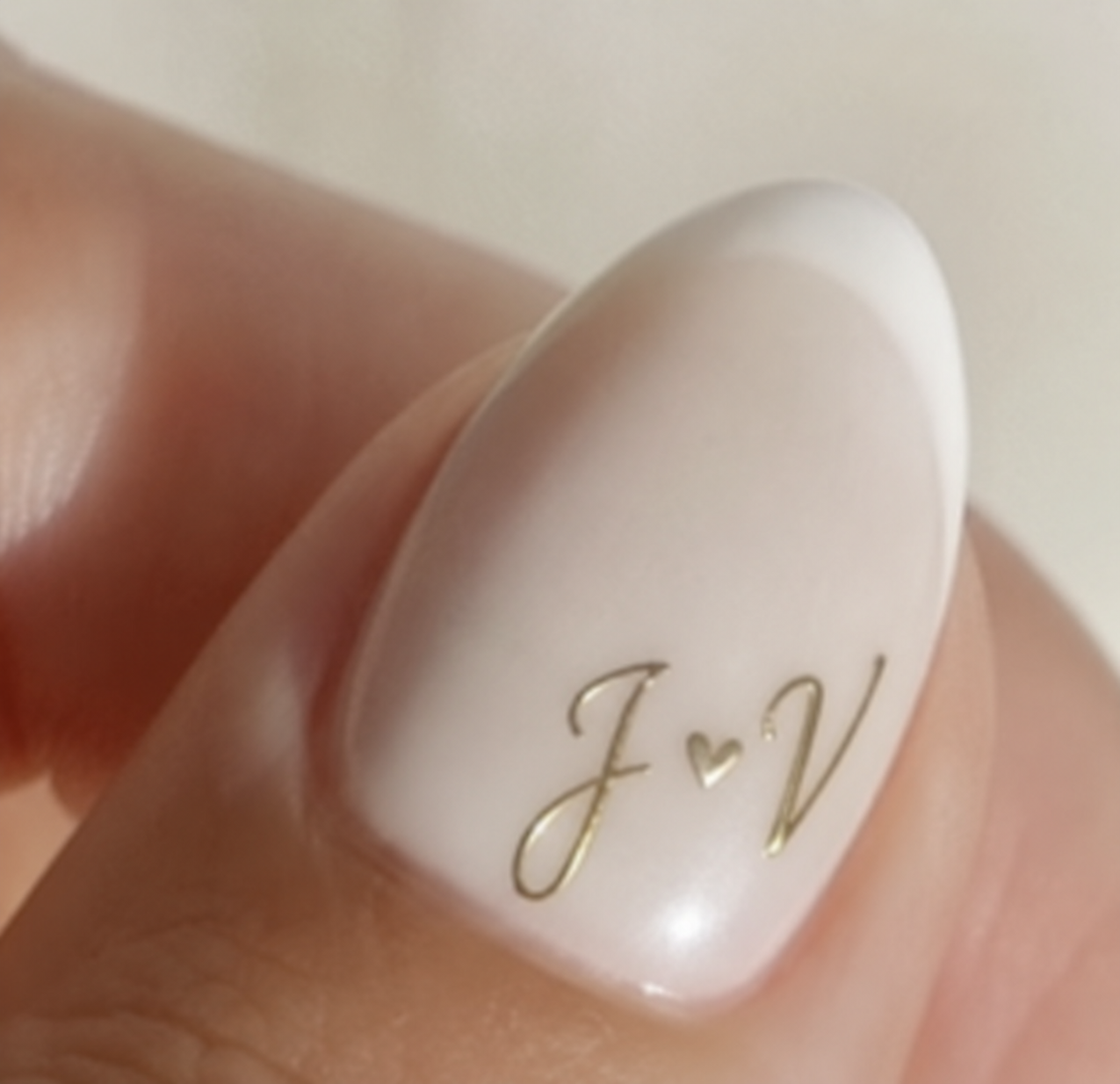 Close-up of an oval-shaped French manicure style nail with the gold initials 'J' and 'V' separated by a small heart.