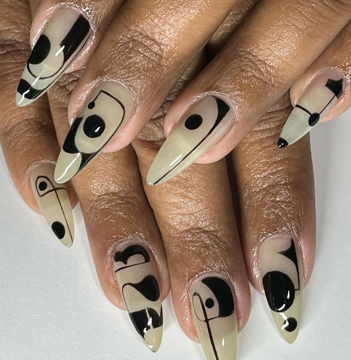 Close up of manicured hands with long, pointed nails decorated with black abstract lines and dots on a light beige base.