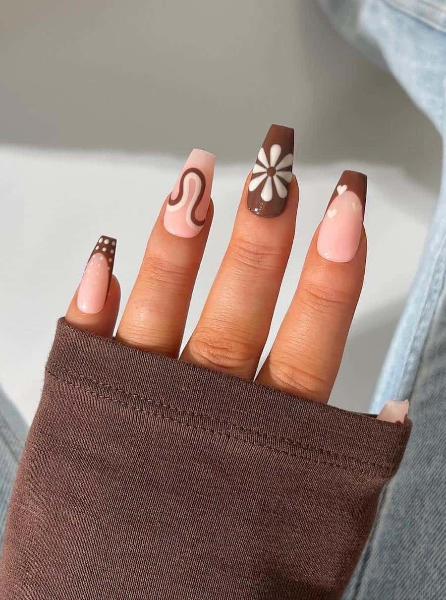 Hand with coffin-shaped nails featuring designs including a swirl, a daisy, polka dots, and a heart, in shades of brown and…