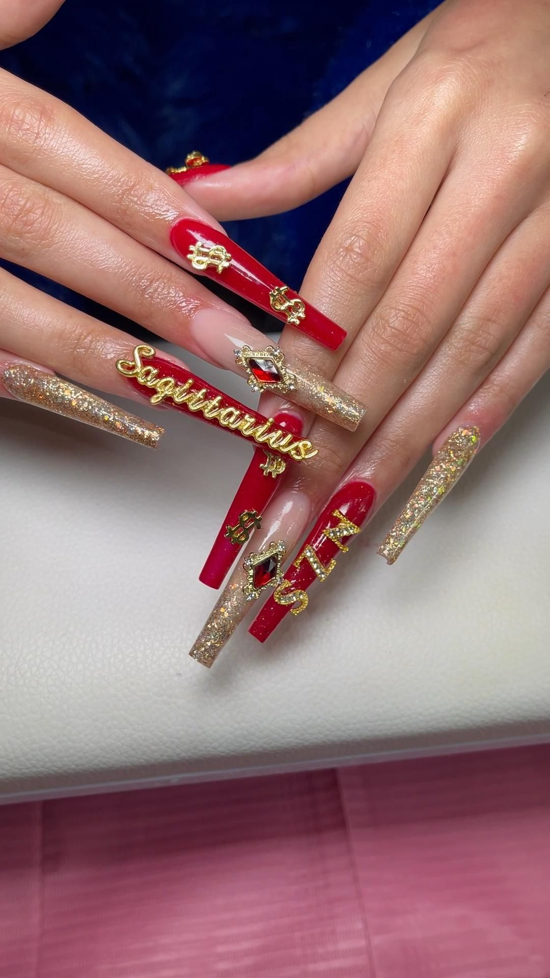 Long artificial red and gold glitter nails with dollar signs, jewels, and "Sagittarius" written in gold lettering.
