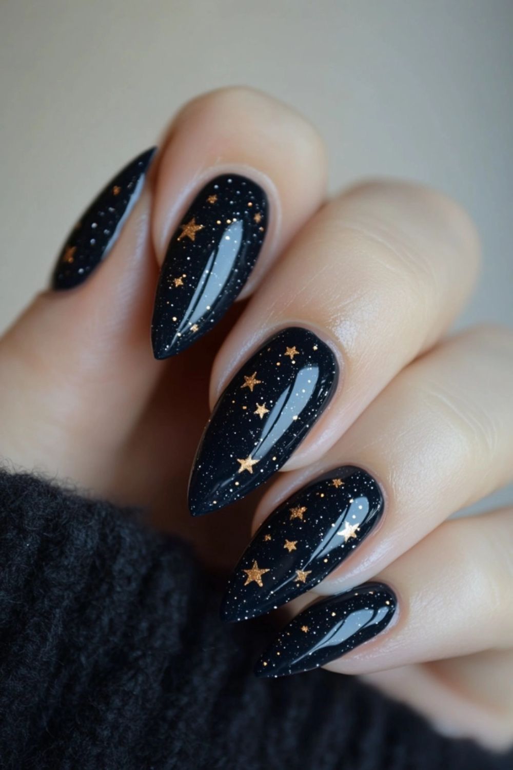 Pointed almond-shaped nails in black with glitter and gold star decals, shown against a dark sweater.