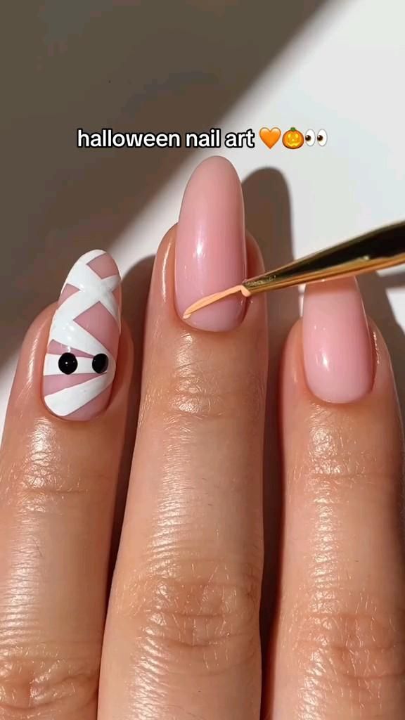 Close-up of a hand with one nail painted to look like a mummy, the others have a light pink base coat being applied.