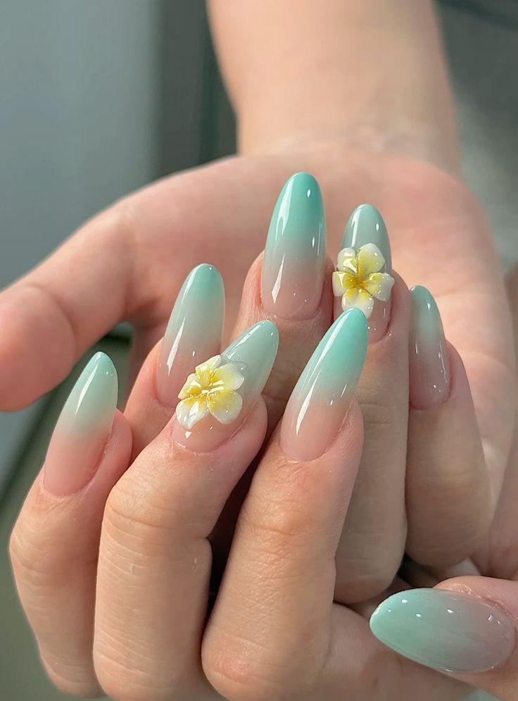 Close-up of a hand with long, oval-shaped nails featuring an aqua ombré design and small, raised white and yellow flowers on…