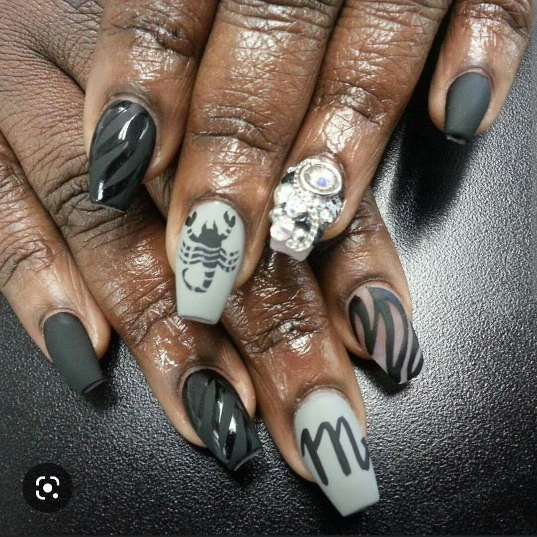 Dark-skinned hand with coffin-shaped acrylic nails. Designs include grey with black scorpion and m-symbol, matte black…