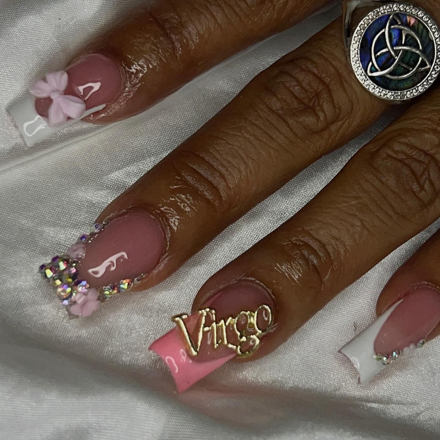Close-up of a hand with long, French-tipped nails featuring a metallic "Virgo" charm, bows, and rhinestones.