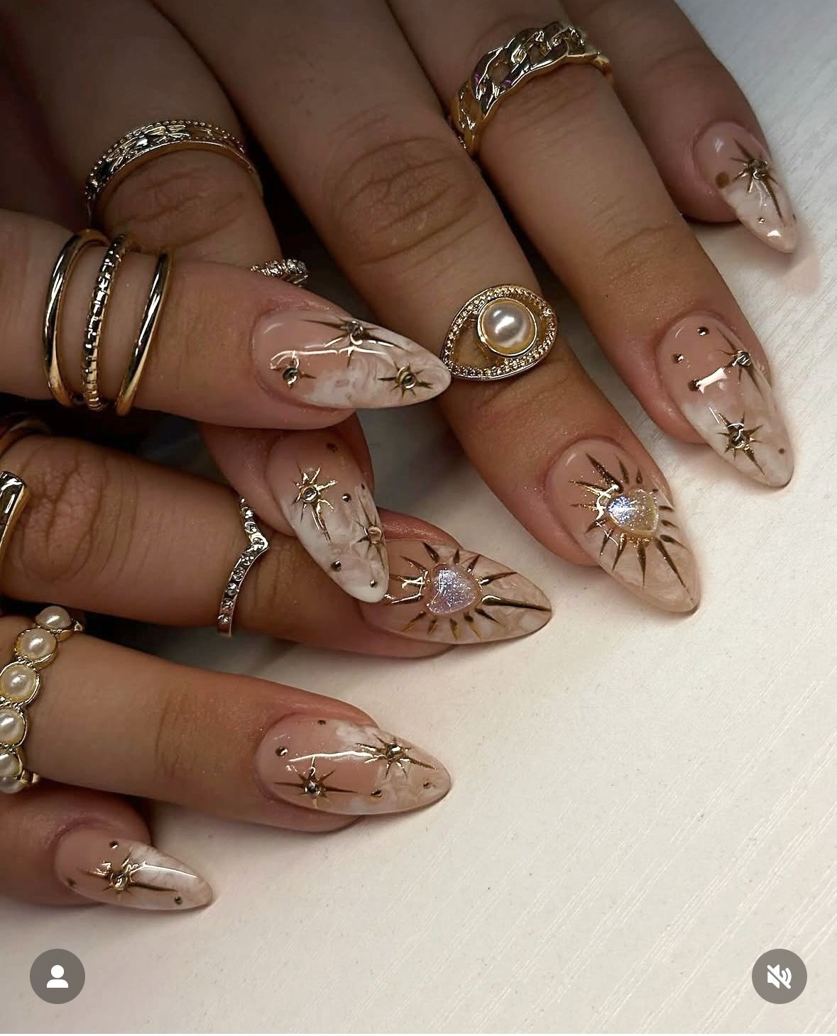 Close-up of manicured hands with long, almond-shaped nails featuring nude and white marble designs, gold starbursts, and…